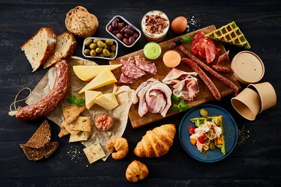 Brunch set oppefra: charcuteri, oste, croissanter, oliven og salat på mørkt bord