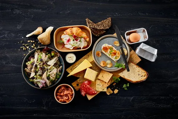 Frokostretter i bokse og skåle på et mørkt bord: sild, salat, smørrebrød og ost