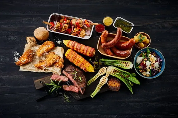 Grillretter set oppefra: pølser, majs, spyd, bøf og salater på mørkt bord